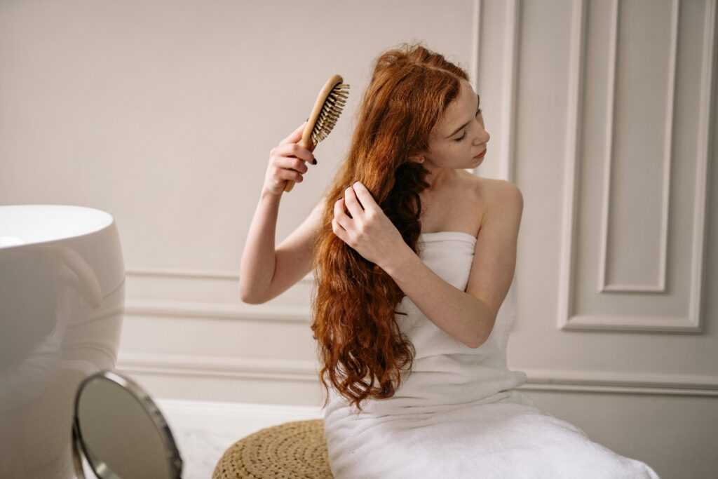 Woman with long red hair wrapped in a towel combing her hair in an elegant bathroom setting.